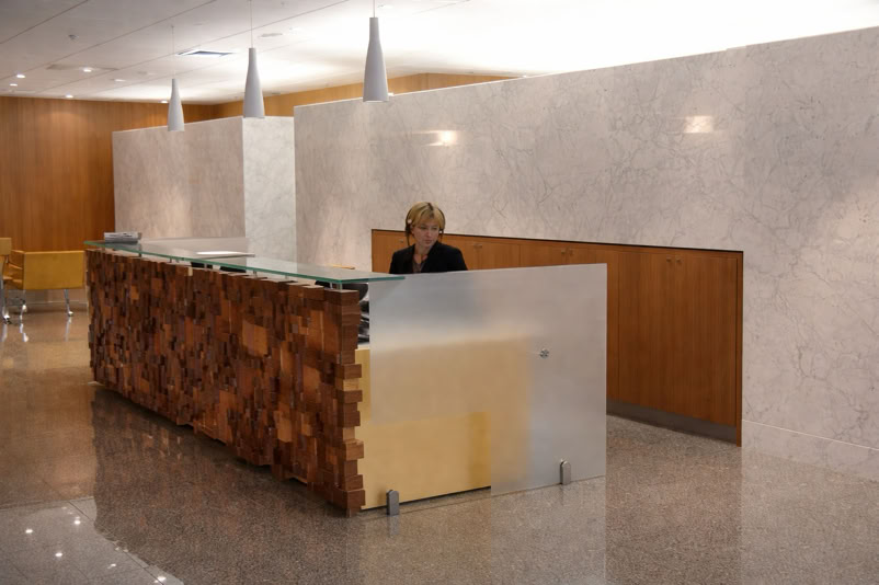A person sitting at a modern reception desk with wood and frosted glass panels, in a lobby with marble walls and a polished floor.
