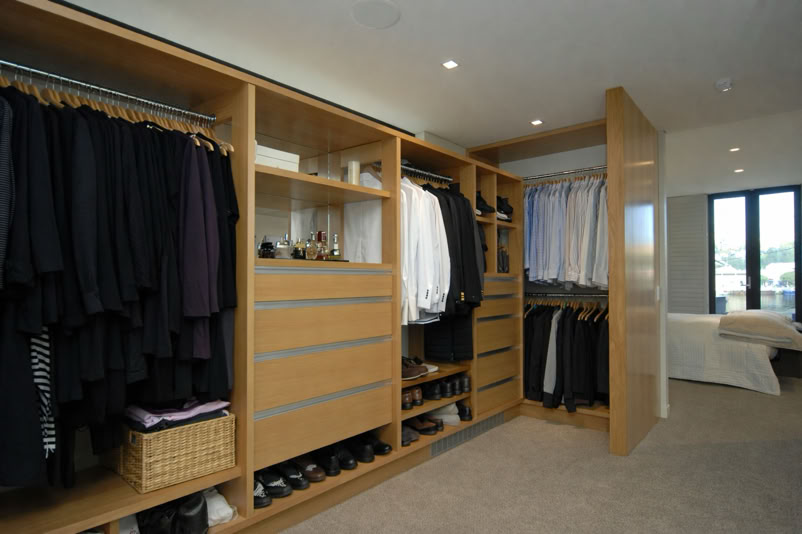 A walk-in closet with wooden shelves and hanging clothes, including shirts and jackets, with drawers and shoe storage below.