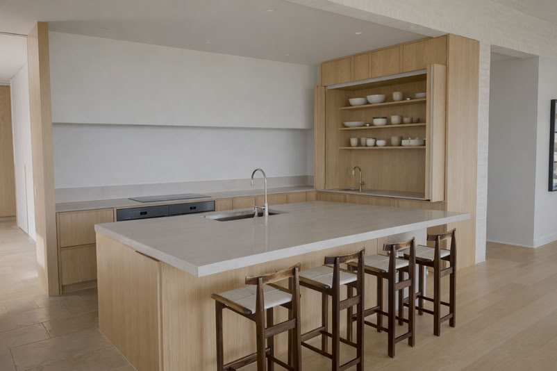 Modern kitchen with a large island, four wooden bar stools, open shelving with dishes, and light wood cabinetry.