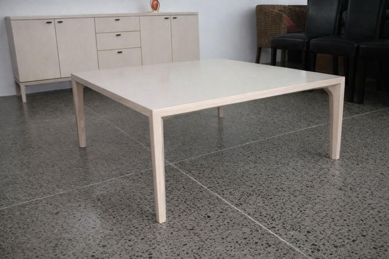 A light-colored square table on a polished concrete floor, with a beige sideboard and black chairs in the background.