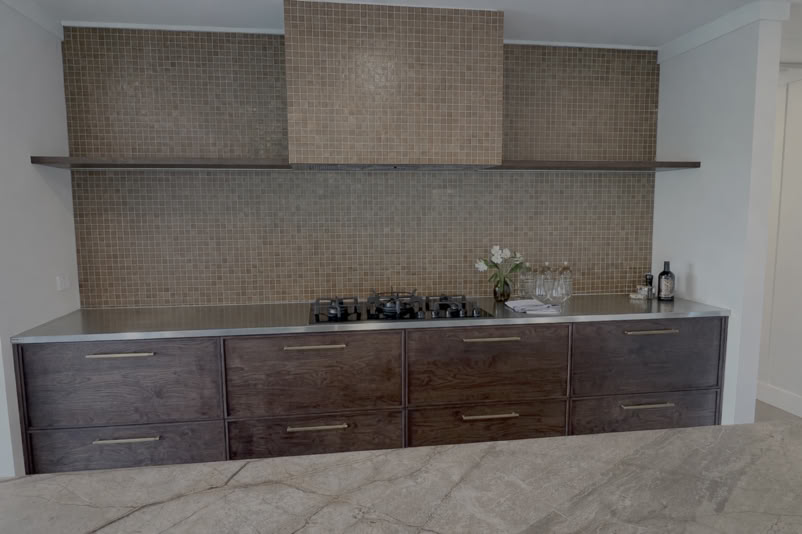 A modern kitchen with dark wooden cabinets, a stainless steel countertop, a gas stove, and a textured backsplash. A shelf holds a vase with flowers and a bottle.