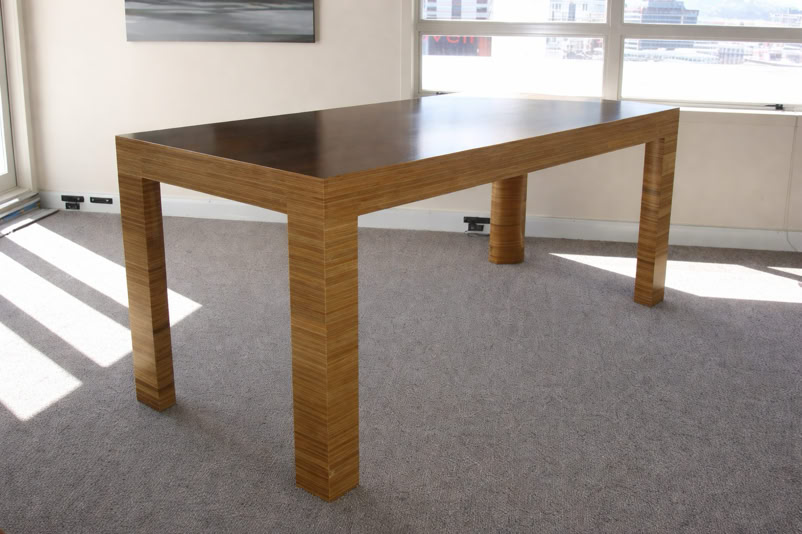 A wooden table with a dark top and light-colored legs, placed on a carpeted floor in a room with large windows.