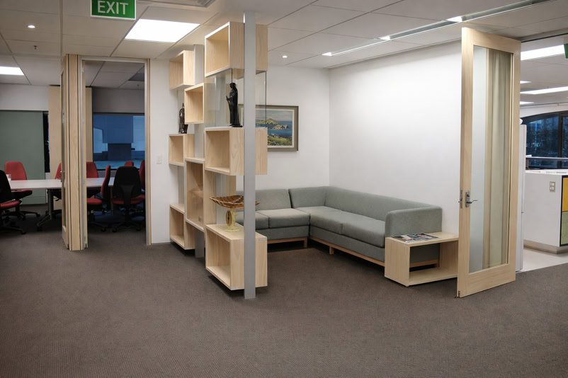 Office space with a gray sectional sofa, wooden shelving unit, open door, and a conference room with red chairs visible in the background.