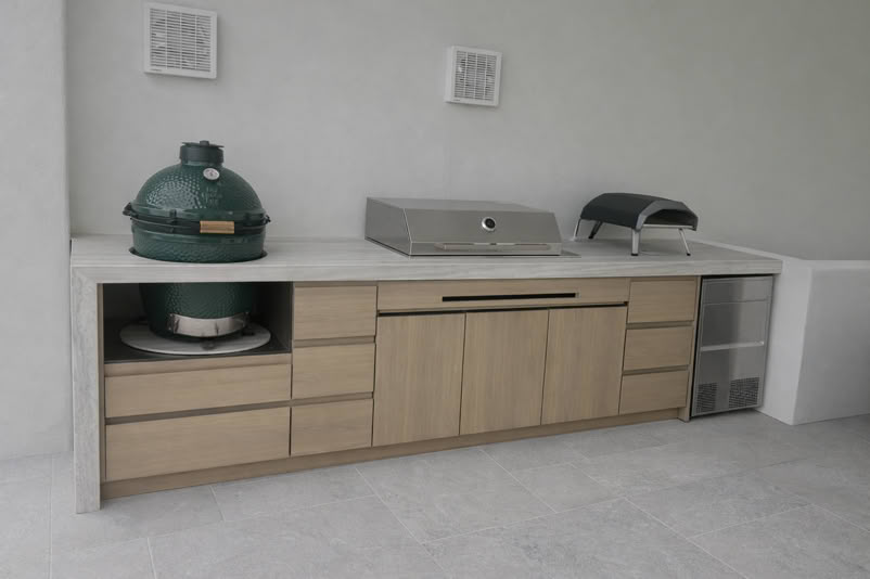 Outdoor kitchen setup with a green ceramic grill, a stainless steel grill, a small pizza oven, and a mini fridge, all set on a wooden counter with drawers and cabinets.