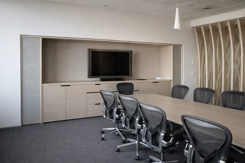 A modern conference room with a long table, several office chairs, a wall-mounted TV, and a wooden cabinet.