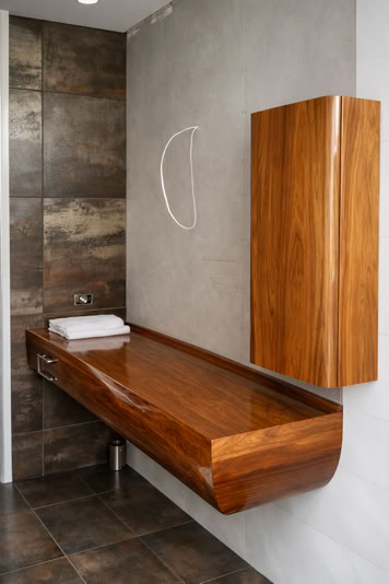 Modern bathroom with a wooden floating vanity, a wall-mounted cabinet, and a towel on the counter. The walls are tiled in a dark, textured pattern.
