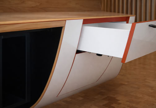 A modern wooden cabinet with a curved design, featuring an open white drawer with orange edges and black handles, set against a wooden floor and wall.