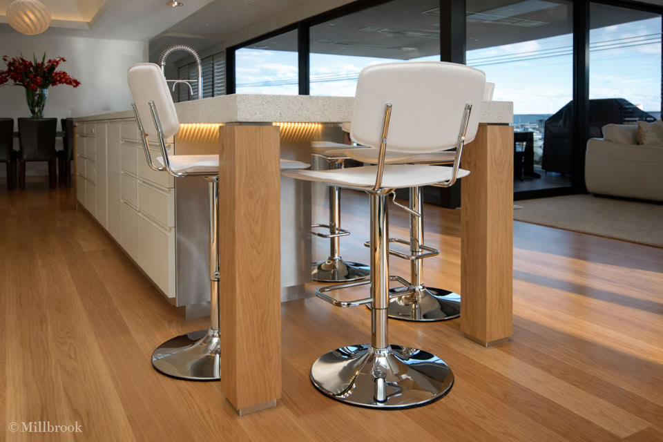 Modern kitchen interior with a wooden floor, white island counter, and two white bar stools with chrome bases. Large windows in the background provide natural light.