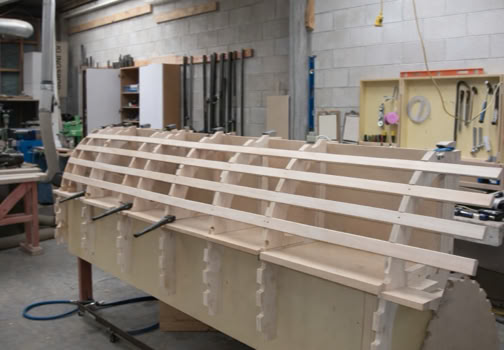 A wooden boat frame under construction in a workshop with tools and equipment in the background.