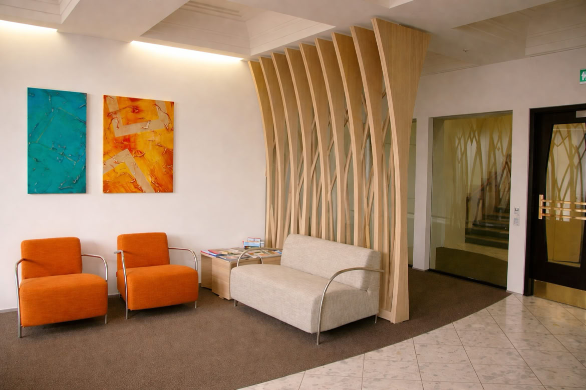 A modern waiting area with two orange chairs and a beige sofa, a wooden partition, two colorful abstract paintings on the wall, and a glass door leading to a staircase.