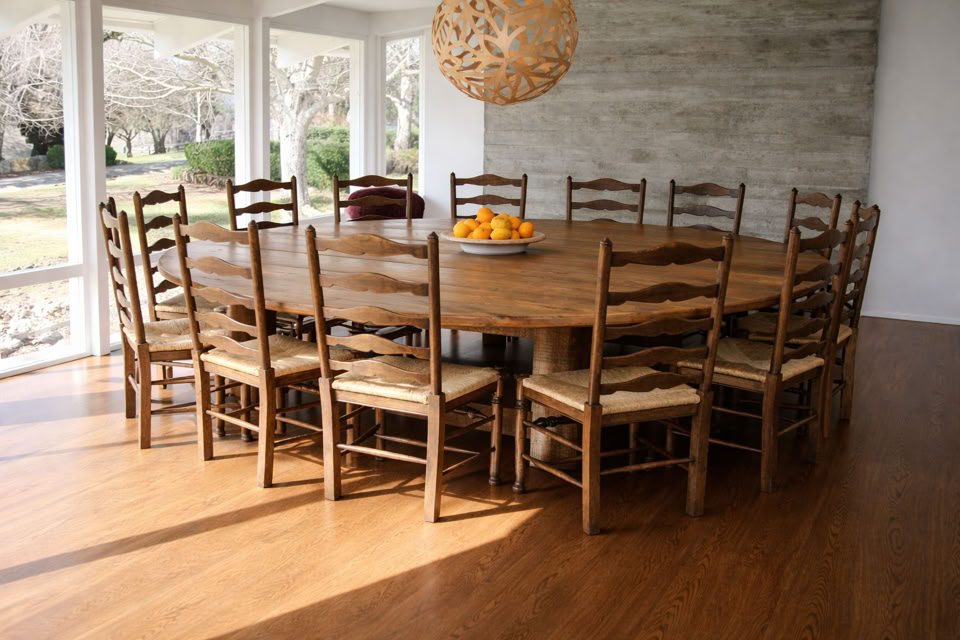 A large round wooden dining table surrounded by twelve wooden chairs with woven seats, placed in a room with large windows and a hanging light fixture above.
