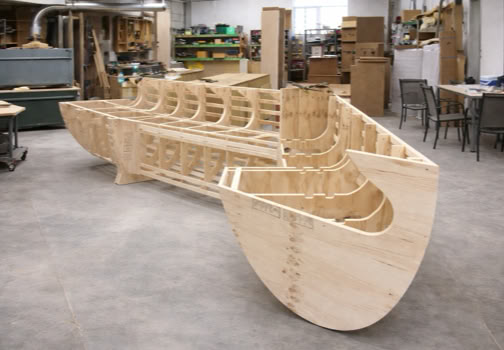 Wooden boat frame under construction in a workshop with various tools and materials in the background.