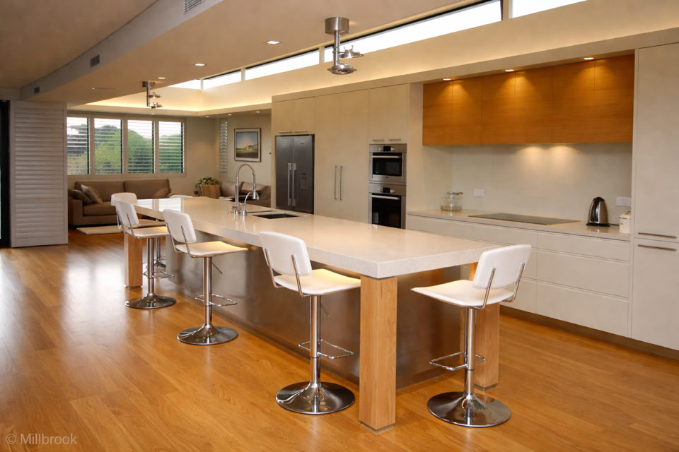 Modern kitchen with a long island, four white bar stools, wooden flooring, built-in appliances, and recessed lighting.