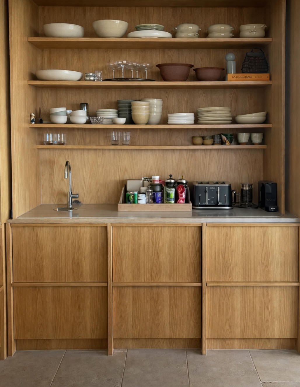 Wooden kitchen shelves with dishes, bowls, and glasses above a countertop with a sink, various bottles, and a toaster.