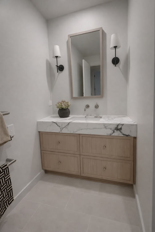 A modern bathroom with a marble countertop, wooden vanity with drawers, a rectangular mirror, two wall-mounted lights, and a small plant on the counter.