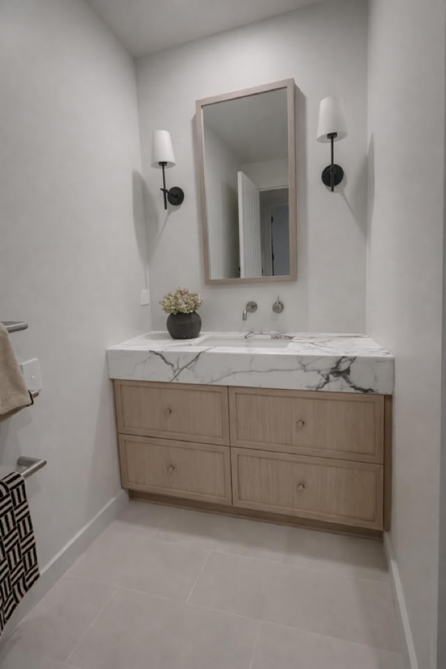 A bathroom with a wooden vanity, marble countertop, rectangular mirror, two wall sconces, and a small plant on the counter.