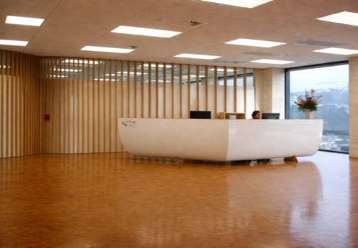 Spacious office lobby with a large white reception desk, wooden flooring, vertical wooden slats on the wall, and a large window with a view outside.