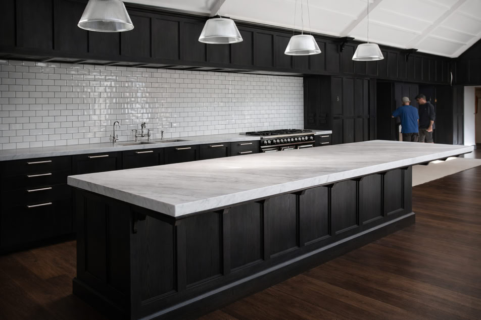 A modern kitchen with a large marble-topped island, dark cabinetry, white subway tile backsplash, and pendant lights. Two people are standing in the background.