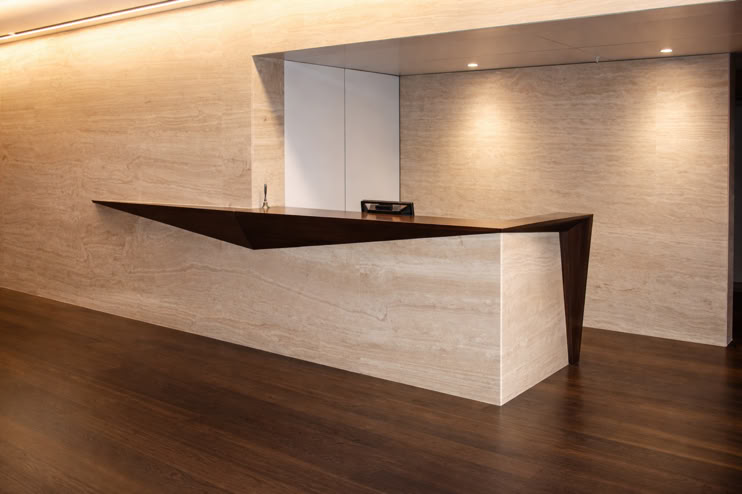 A modern reception desk with a geometric design, featuring a dark wood top and light stone base, set against a beige wall and dark wood floor.