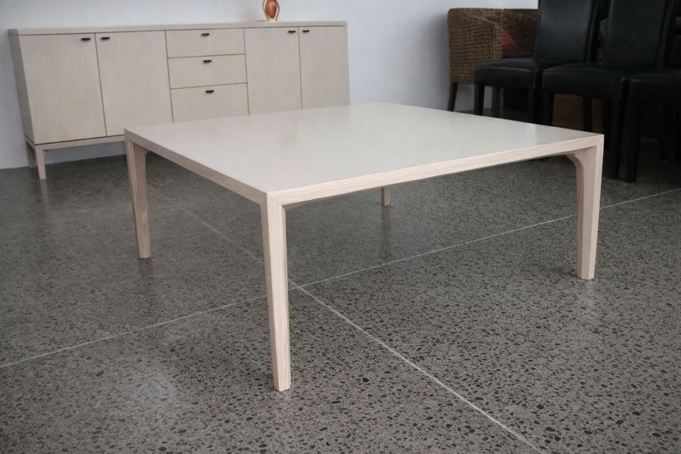 A light-colored, rectangular table on a polished concrete floor, with a beige sideboard and dark chairs in the background.