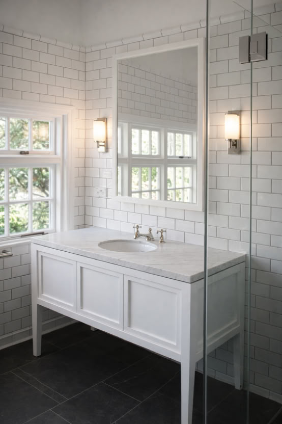 A bathroom with white subway tile walls, a large mirror, a white vanity with a sink, two wall-mounted lights, and a window with a view of greenery.