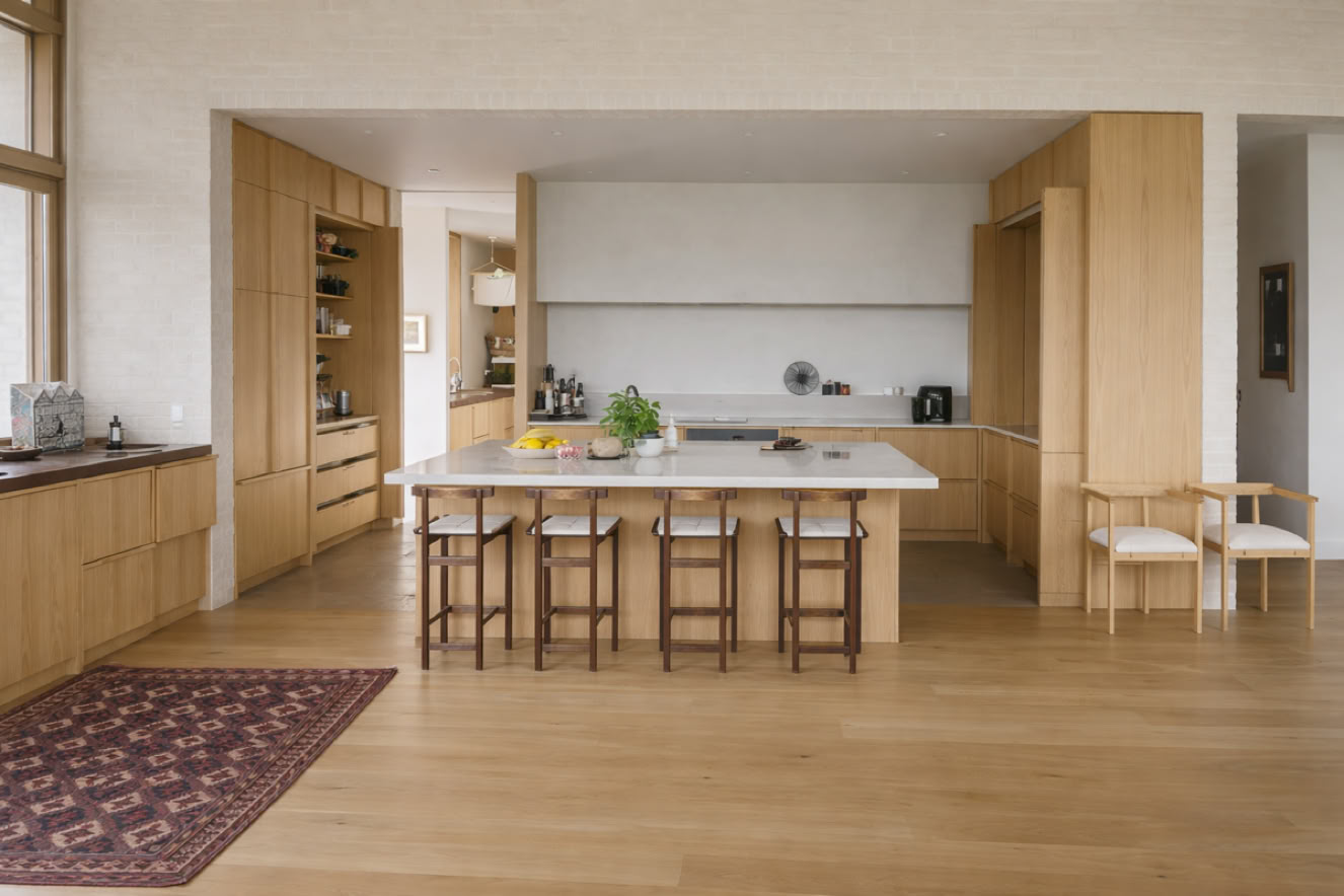 A modern kitchen with wooden cabinets, a large island with stools, a rug on the wooden floor, and a window on the left.