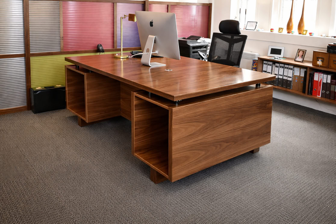 A modern office with a wooden desk, computer monitor, desk lamp, and office chair, surrounded by shelves and storage units.