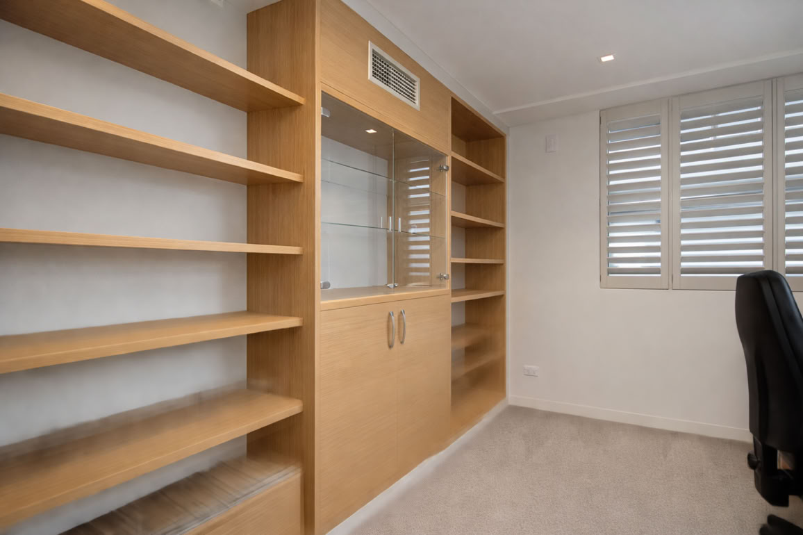 Room with wooden shelves, a cabinet with glass doors, a black office chair, and a window with white shutters.