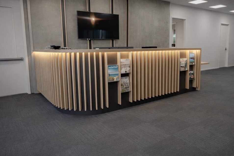 A modern reception desk with vertical wooden slats, illuminated from below, and shelves containing books or magazines. A wall-mounted TV is above the desk, and the room has a gray carpet and white walls.