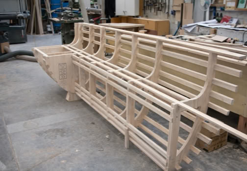 Wooden boat frame under construction in a workshop with various tools and materials in the background.