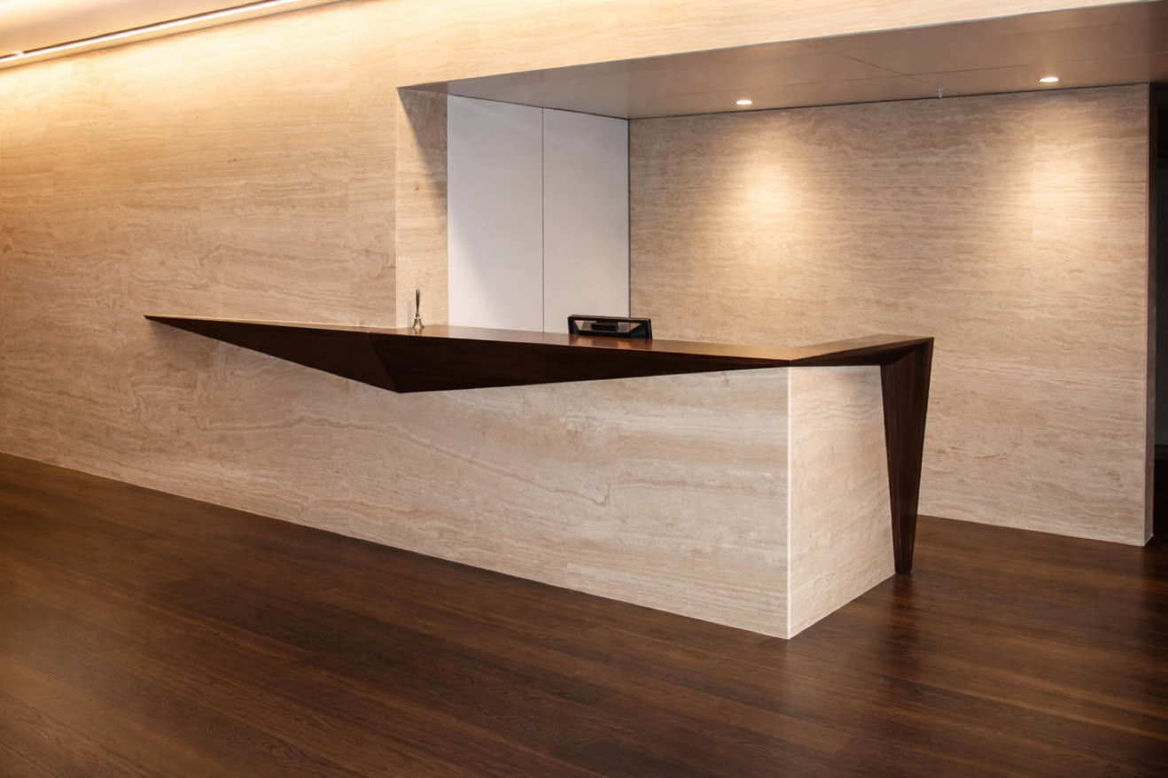 Modern reception desk with a geometric design, featuring a dark wooden top and light stone base, set against a beige wall with recessed lighting.
