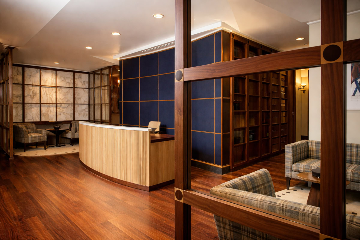 A modern office reception area with a wooden desk, blue accent wall, wooden flooring, and seating area with plaid armchairs.