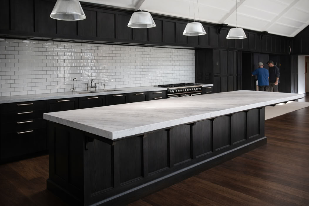 A modern kitchen with a large marble-topped island, dark cabinetry, white subway tile backsplash, and pendant lights. Two people are standing in the background.