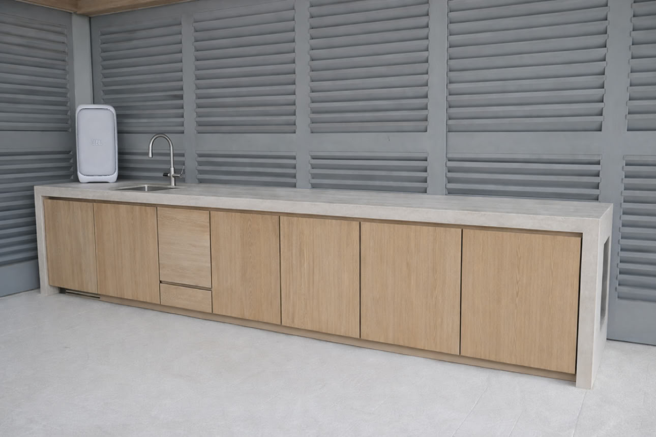 Outdoor kitchen with a long countertop, wooden cabinets, a sink, and a water dispenser against a gray slatted wall.