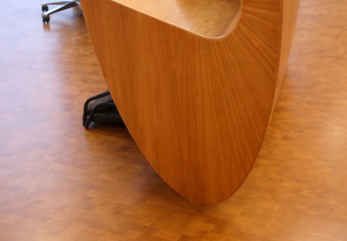 Modern wooden desk with a curved design, featuring a computer monitor, keyboard, and office supplies on top, with a red chair nearby on a wooden floor.