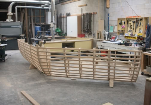 Wooden boat frame under construction in a workshop with tools and equipment in the background.