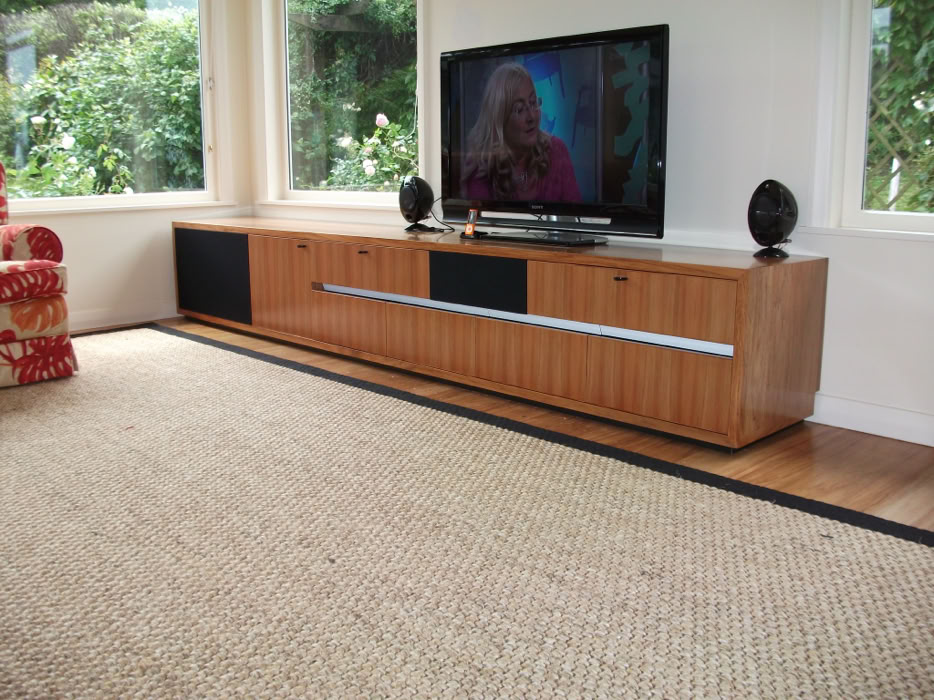 A living room with a large wooden entertainment unit, a flat-screen TV, two black speakers, and a woven rug on the floor.