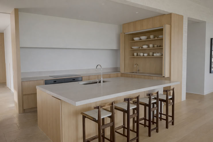 Modern kitchen with a large island, four wooden bar stools, open shelving with dishes, and light wood cabinetry.