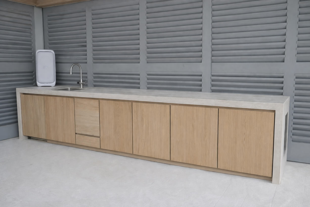 Outdoor kitchen with a long countertop, wooden cabinets, a sink, and a small refrigerator, set against a gray slatted wall.