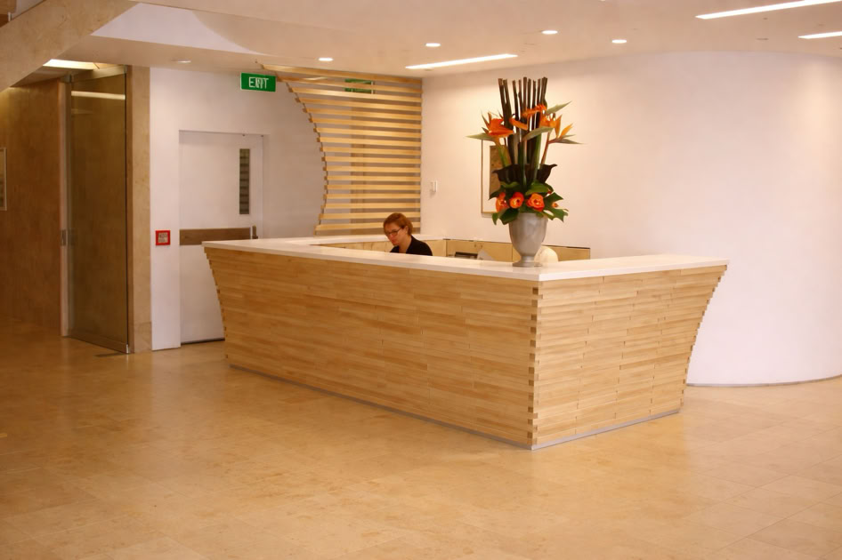 A modern reception area with a wooden desk, a person seated behind it, and a large vase with flowers on the desk. The background features a white wall and a wooden door.