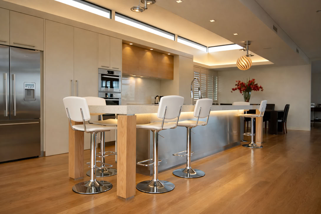 Modern kitchen with a long island, four white bar stools, stainless steel appliances, wooden floor, and a dining area with a table and chairs in the background.