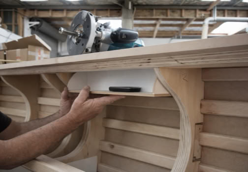 Person installing a white device under a wooden structure with curved supports, in a workshop setting.