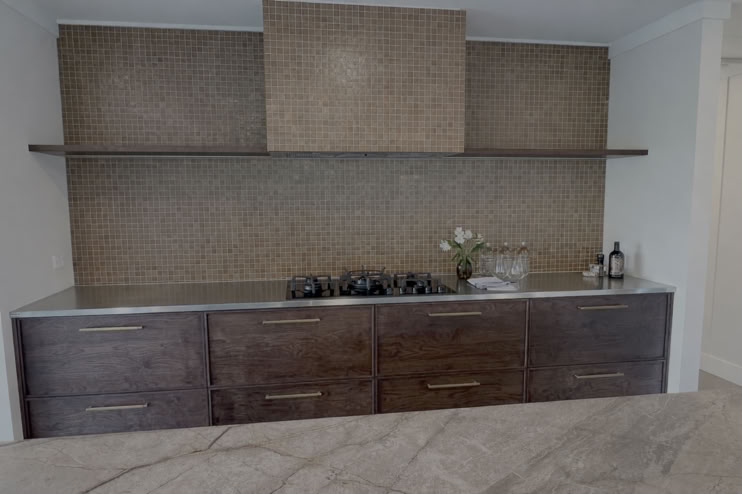 A modern kitchen with dark wooden cabinets, a stainless steel countertop, a gas stove, and a textured backsplash. A shelf holds a vase with flowers and a bottle.