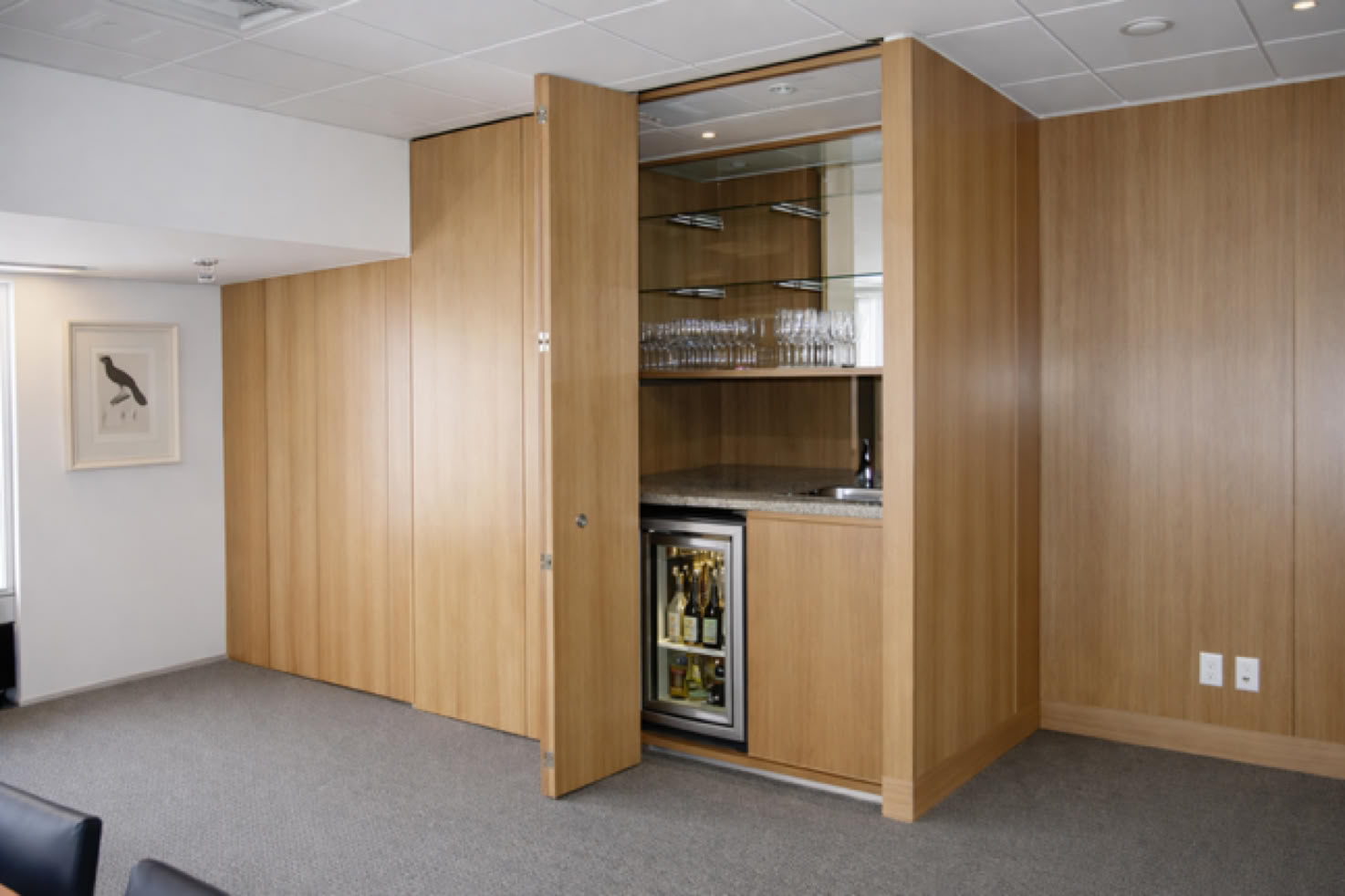Wood-paneled room with an open cabinet revealing glass shelves and a mini fridge, a framed bird illustration on the wall, and a gray carpeted floor.