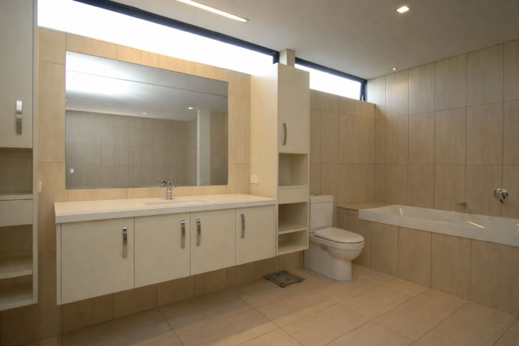Modern bathroom with a large mirror above a double sink vanity, a toilet, and a bathtub, all set against beige tiled walls and floor.